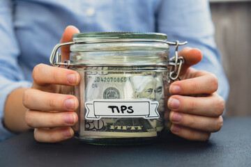 Female hands Saving Money In Glass Jar filled with Dollars banknotes. TIPS transcription in front...