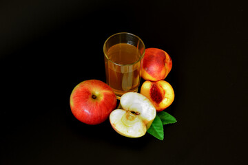 A glass of freshly squeezed fruit juice on a black background, next to pieces of ripe peach and red apple.