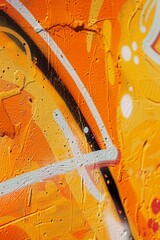 A close-up view of a wall reveals graffiti in vibrant orange and yellow colors, showcasing texture detail and paint splatter in a street art style.