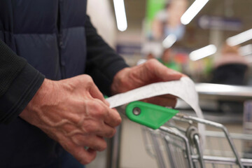 The hands of an unrecognizable older man hold a sales receipt next to a grocery cart in a supermarket.