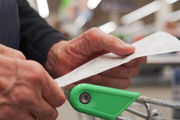The hands of an unrecognizable older man hold a sales receipt next to a grocery cart in a...