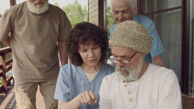 Medium close-up shot of elderly male and female Caucasian retirement community residents and smiling nurse in uniform posing together on terrace, taking fun selfies on smartphone then checking result