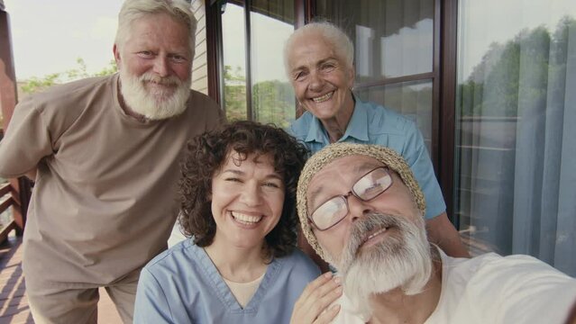 Medium close-up UGC footage of senior male and female Caucasian assisted living home residents and nurse in uniform taking fun photo or video selfies together on smartphone and talking on camera