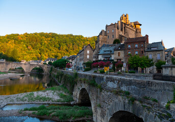 Summer view of scenic French township of Estaing on bank of Lot River overlooking medieval stone...