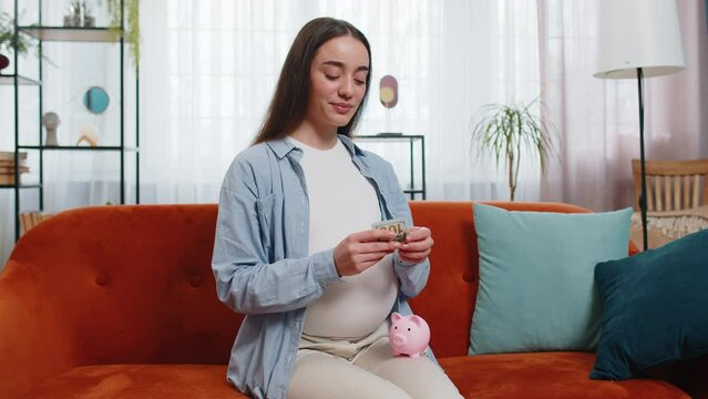 Happy thinking young pregnant woman inserting banknote in piggybank and caressing big belly sitting on sofa in living room at home. Smiling future mother lady on couch saving for baby's future concept