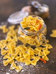 Glass jar filled with dry farfalle pasta and scattered around on dark wooden background.