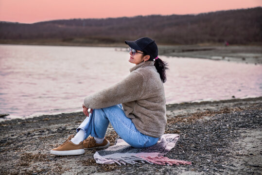 49 year old Russian woman in jacket sitting on a beach and thinking against red sunset
