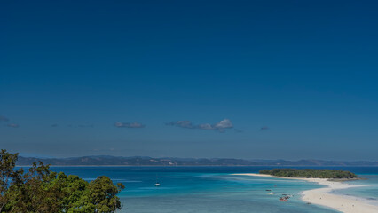 A delightful tropical landscape. A winding sandbar leads to a small island covered with green vegetation. The boats are anchored in the aquamarine ocean. Mountains against the blue sky in the distance