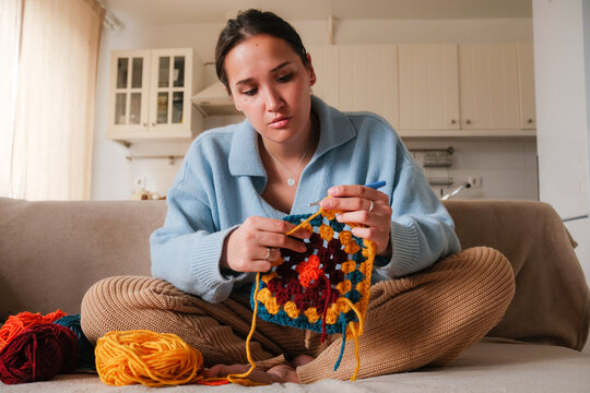 Young woman knitting crocheting with colored yarn granny square while watching online tutorial on phone at home. Woman watching needlework lessons, home hobbies