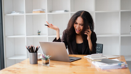 Businesswoman showing frustration during a phone call, with a laptop and documents on her office...