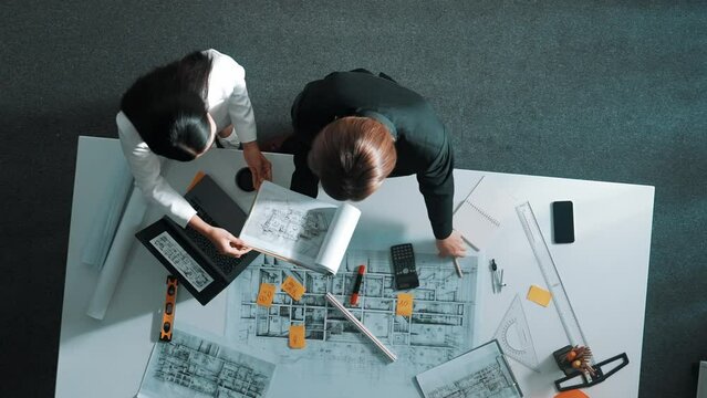 Top view of skilled project manager holding project plan and explain house design to architect engineer at meeting table with equipment, ruler, safety helmet and laptop scatter around. Alimentation.
