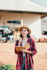 Obraz premium Smiling farmer woman stands holding a notebook, dressed in a straw hat and plaid shirt, with a tractor in the background.