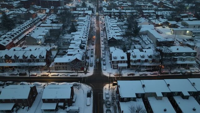 Twilight over snow-blanketed American city street, lined with row houses and lit by warm glows. Aerial reverse dolly in quaint neighborhood suburb.