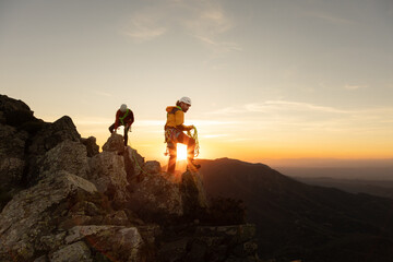 Two people are climbing a mountain, one of them wearing a yellow jacket. The sun is setting in the...