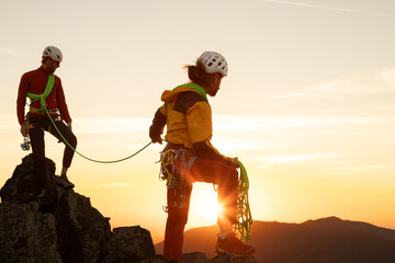 Two people are on a mountain, one of them is wearing a yellow jacket. The sun is setting in the...