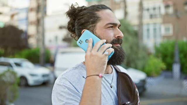 Handsome hispanic man with a beard and manbun hairstyle talking on a smartphone on an urban city street during daytime.