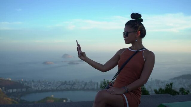 A woman takes a selfie near Christ the Redeemer, capturing the backdrop of Rio de Janeiro city and distant islands.