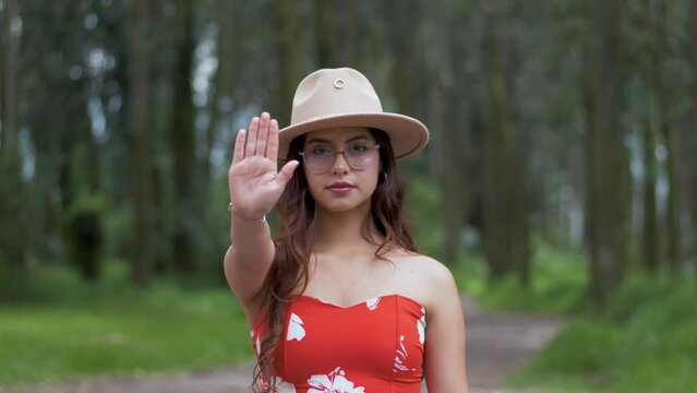 Young woman in a red summer dress and hat raises her hand as a stop sign. Cease and desist, it's not okay to continue. Static slow motion in nature