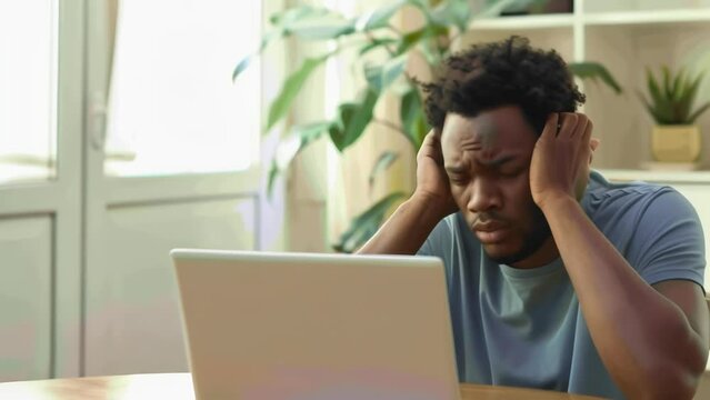 A man sitting at a laptop, head in hands, appears dejected and stressed, his expression gloomy. Houseplants and a window behind him suggest an indoor setting. 