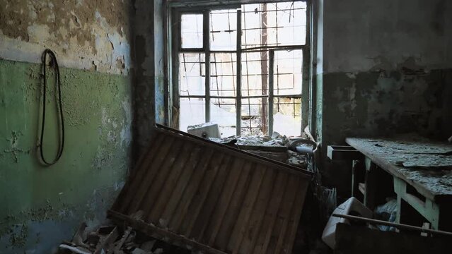 An eerie and deserted scene of an abandoned building. The room has a broken window, broken furniture and peeling paint on walls. There is rubbish scattered on floor, an atmosphere of decay