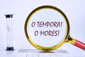 o tempora, o mores (O, the times O, the morals) Latin phrase through a magnifying glass on a gray background next to an hourglass - obrazy, fototapety, plakaty