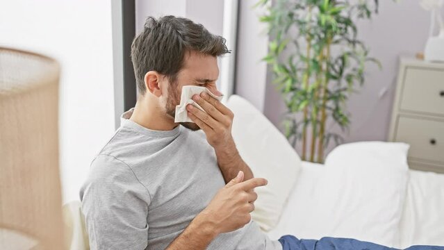 Sick young man with a beard blowing his nose while sitting in a bedroom, depicting illness and homecare.