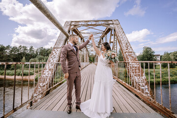 Valmiera, Latvia- July 28, 2024 - Bride and groom dance on a rustic bridge, the groom holding the...