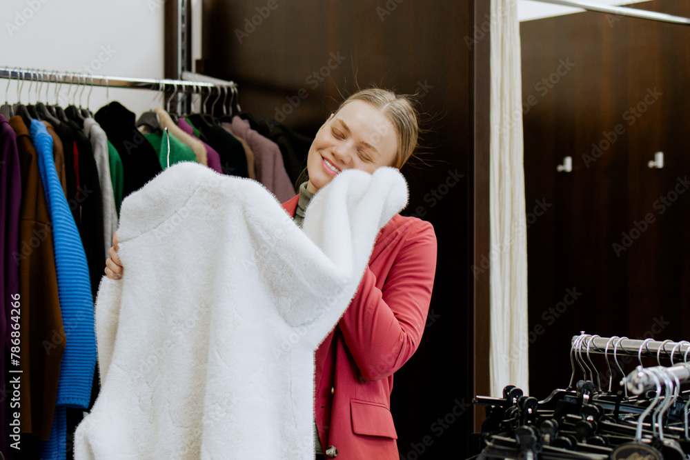 Wall mural Happy woman touches a white faux fur coat and tries it on for herself, in a women's clothing store, in a boutique. Shopping in a clothing store.