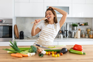 Excited woman singing and dancing in modern kitchen at home, happy woman holding spatula as microphone, dancing, listening to music, having fun with kitchenware, preparing breakfast