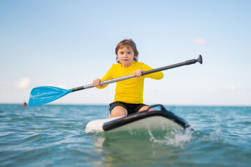Kid riding on a paddle board. Summer holidays. Kid paddle surf surfer little blonde boy in summer beach. Happy Child floating on a paddleboard in the ocean. Kid doing paddle in summer. - 786865839