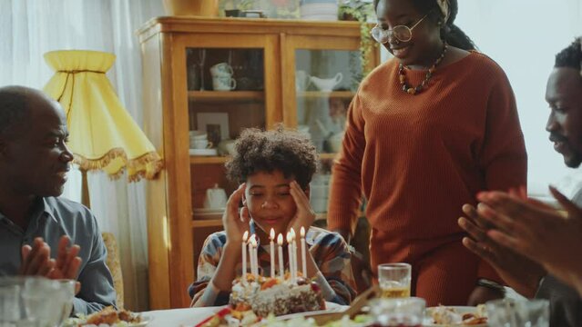 Little African American boy covering his eyes, then blowing candles on cake and smiling while family clapping hands and greeting him on Birthday dinner party at home
