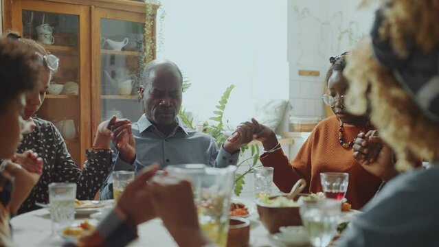 Big African American family sitting around the dinner table, holding hands and praying with eyes closed during holiday celebration at home