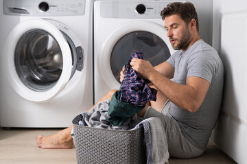 Middle aged single man with laundry basket and dirty clothes near washing machine at home. Millennial man putting clothes into washing machine. Displeased hispanic man wash clothes in washing machine. - 786879418