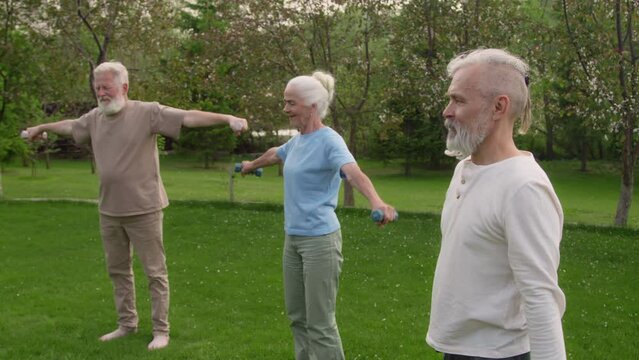 Medium full footage of three senior Caucasian retirement home residents in casual clothes standing in garden, doing weight training exercises with dumbbells, for maintaining fitness and health