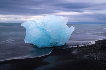 t&uuml;rkiser Eisbrocken (kleiner Eisberg) liegt auf schwarzem Lavastrand, Naturidyll Diamondbeach in Island, pastellfarbene Bew&ouml;lkung am fr&uuml;hen Morgen