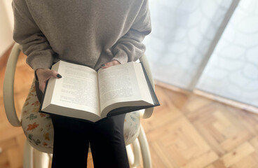 Fototapeta premium Beautiful woman reading a book in the living room of her house with a large window behind it, book day, entertainment, exercising the mind