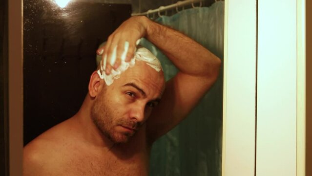 moody shot of a bald bearded man, applying shaving foam to his head, preparing it for a shave, in front of the bathroom mirror. The man is caucasian and tan with a hairy chest and arms.
