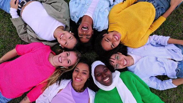 Diverse happy woman bonding together at city park. Multiracial group of female friends lying in circle on the grass, enjoying time together looking at camera. Female friendship concept.