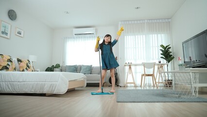 Attractive cute daughter mopping floor while moving to music at modern house. Energetic girl with cleaning gloves doing housework and enjoy cleaning bedroom while singing a songs. Lifestyle. Pedagogy.