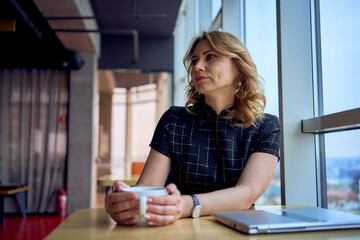 stunning mature woman at a table in a cafe drinking coffee near panoramic windows with a view of the city