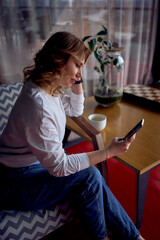 cute middle age woman search on her phone in a beautiful cafe with greenery and light panels in the background