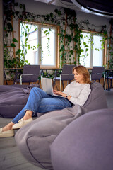 Woman  on a puff chair  working on her laptop in coworking with modern interior design,  neutral...