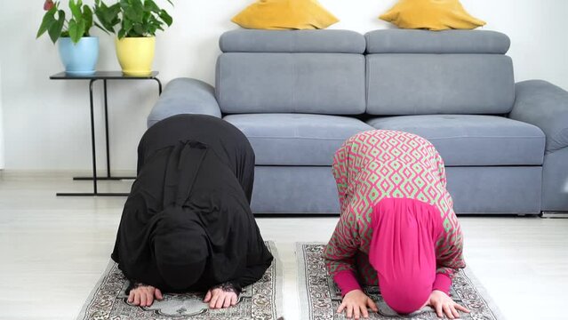 caucasian Muslim girls Praying Sujud at home. A woman in a traditional black abaya and hijab and a girl in a pink dress pray Salat. Mother and daughter read Namaz.