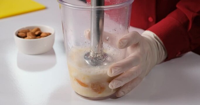 White gloved hands holding a transparent blender with apricots and almonds inside. Immersion blender to blend the contents.