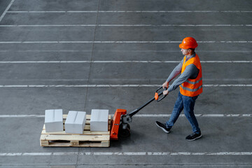 Worker wearing protective helmet, work wear, vest, carrying pallet truck with boxes in warehouse