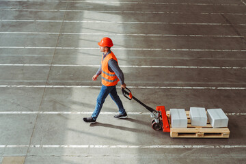 Factory worker, engineer wearing hard hat, vest, work clothes carrying pallet truck with boxes - obrazy, fototapety, plakaty