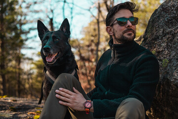 Young white man with sunglasses sitting with his black dog in the forest..jpg