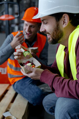 Bearded, smiling worker wearing hard hat and vest holding lunch box, eating food, during break