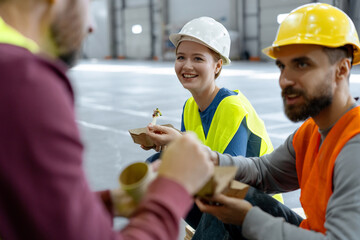 Smiling workers, construction wearing hard hats, vests sitting together, eating lunch, talking