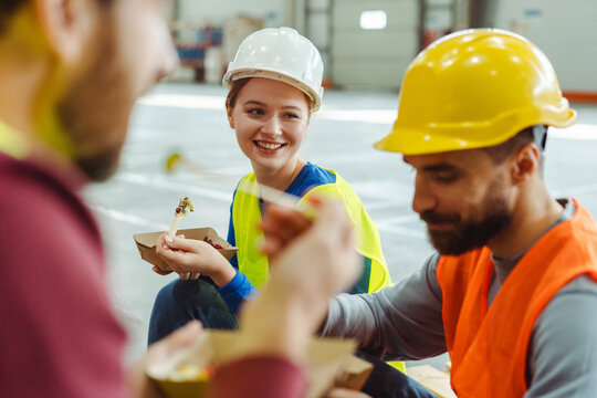 Group of workers, managers, wearing hard hats and vests having lunch, eating, smiling, talking